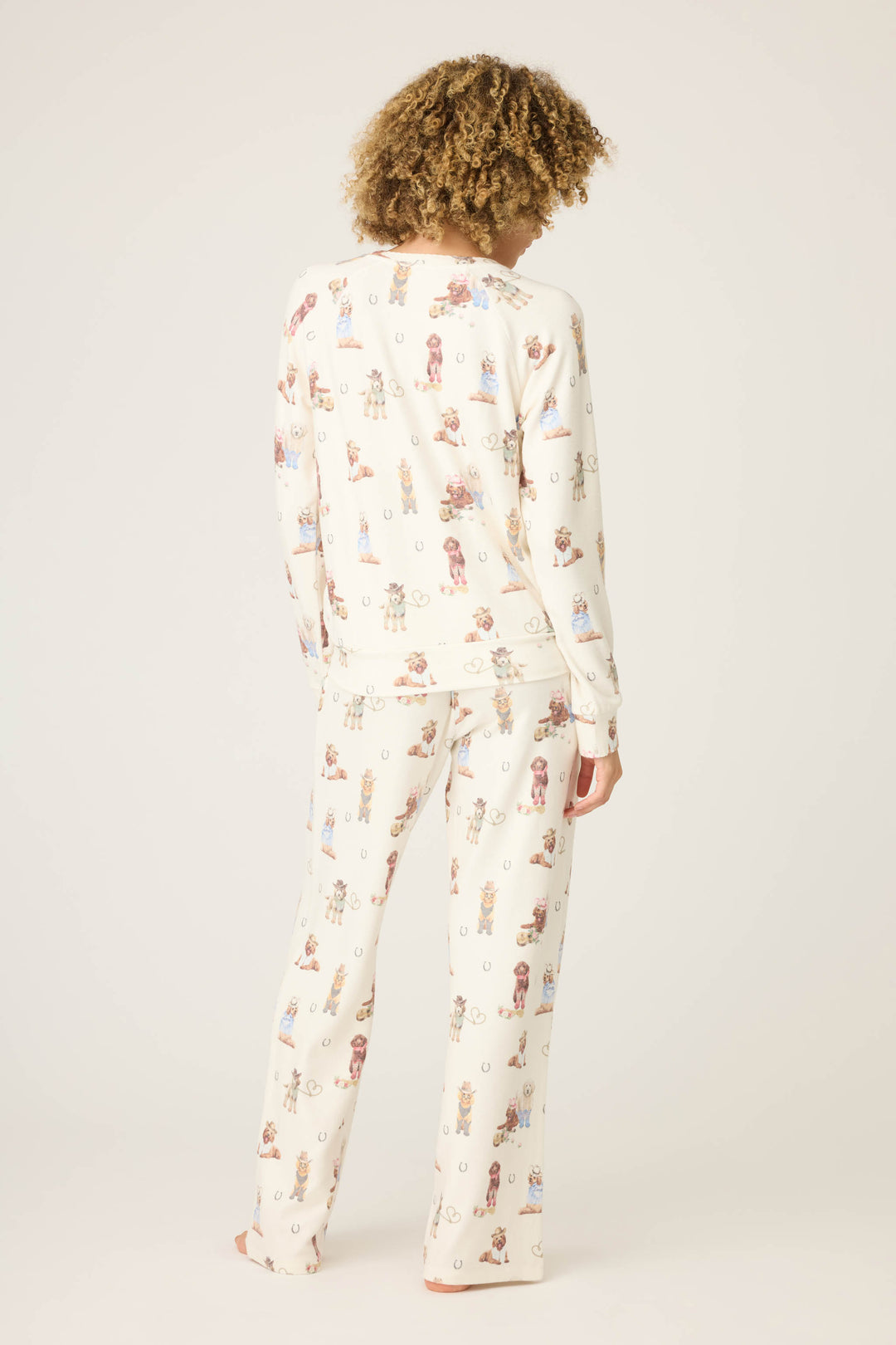 A person with curly hair stands barefoot, facing away from the camera, wearing the cream-colored DOGS PJ SET RUFF ON THE RANGE by P.J. Salvage with a playful small dog print, set against a plain light background.