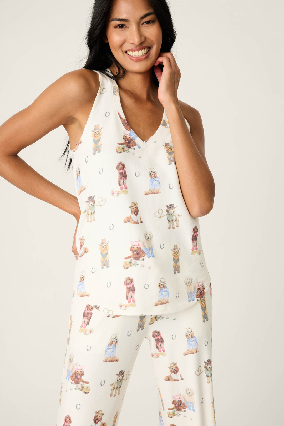 A woman smiles in the P.J. Salvage DOGS V-Neck Tank Ruff on the Range, a matching pajama set with a white background, colorful dog prints, and horseshoe motifs. One hand rests on her hip, the other touches her face.