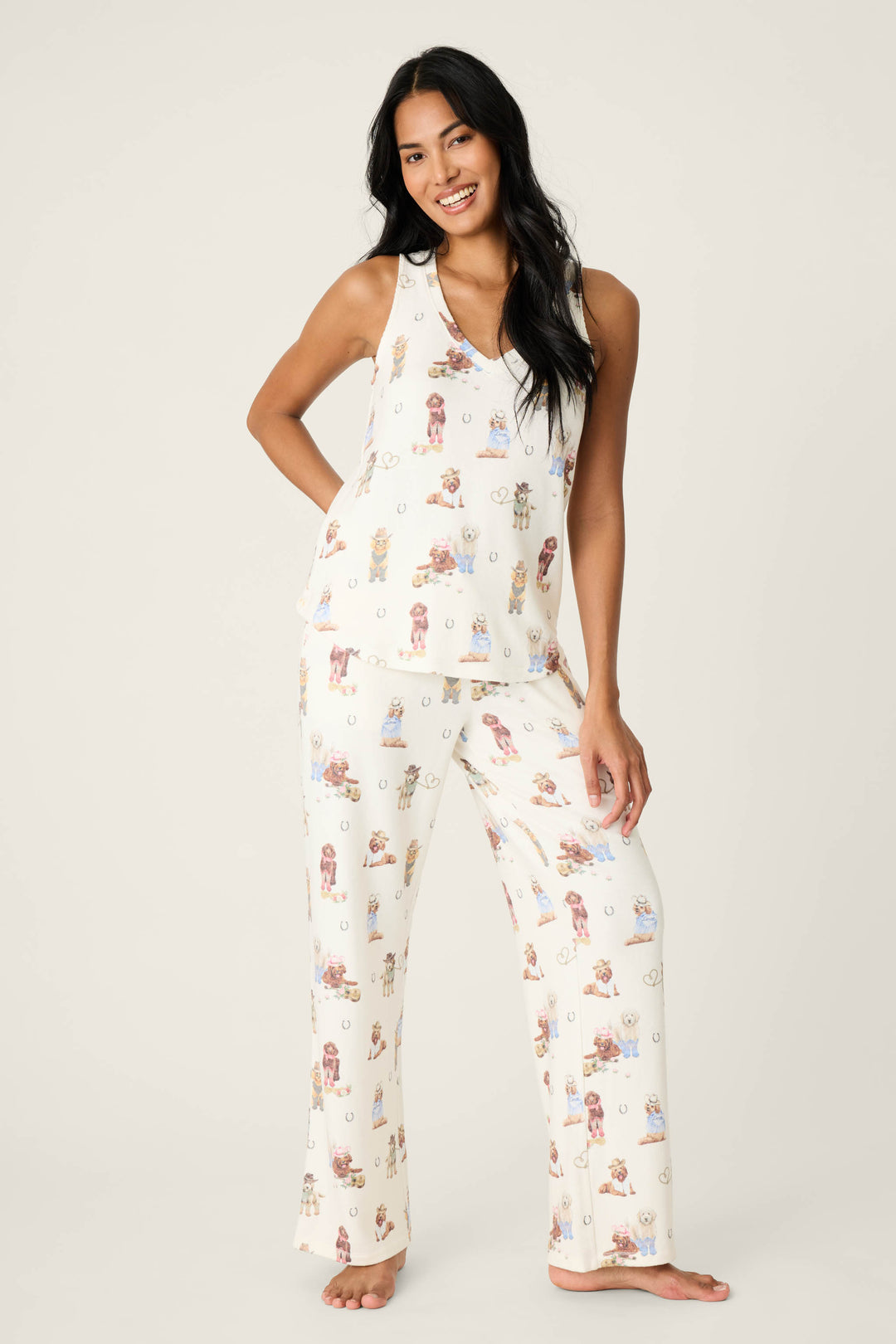 A woman stands barefoot against a plain background, smiling and wearing the P.J. Salvage DOGS V-Neck Tank Ruff On The Range cream pajama set with a sleeveless top and long pants featuring small animal prints.
