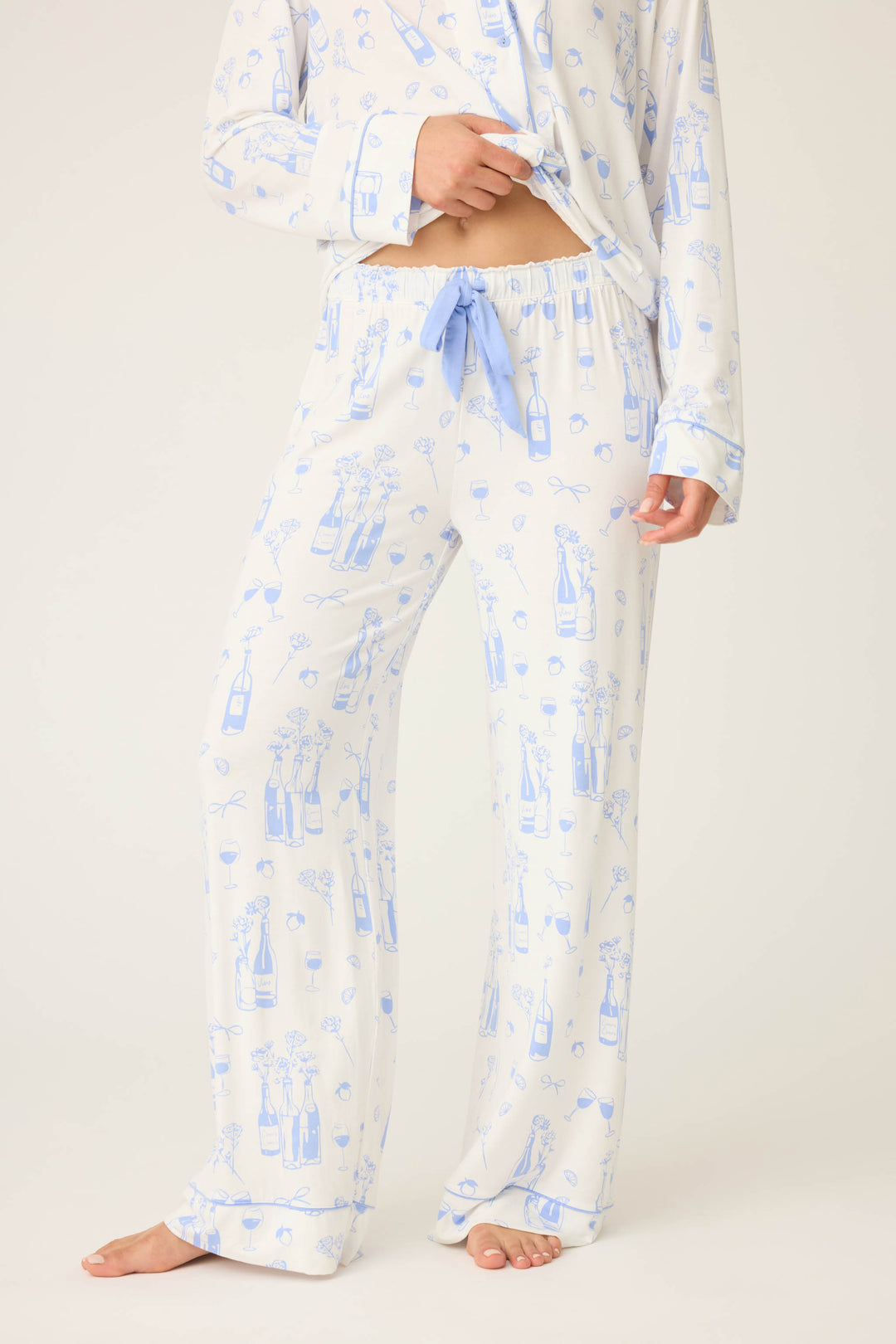 A person wears the P.J. Salvage Parisian Dreams Whimsical Print Open Leg Pant—light-colored pajama pants with a blue bottles-and-glasses pattern and a blue waist drawstring—paired with a matching long-sleeve shirt; their feet are bare.