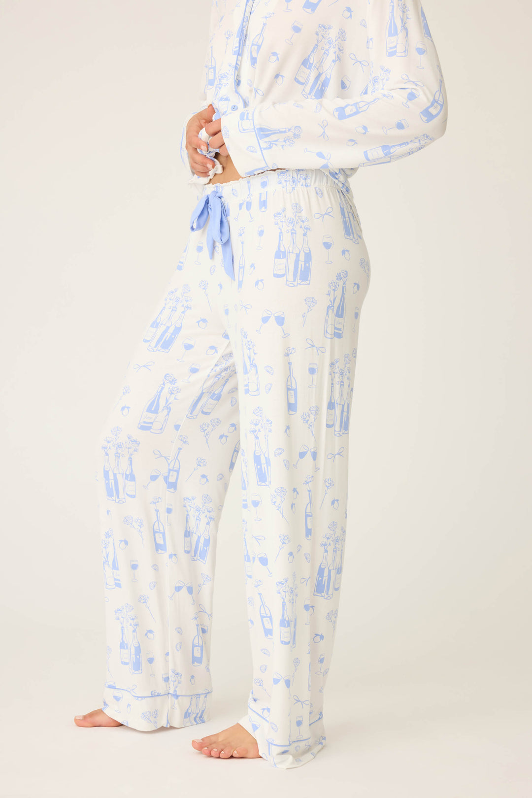 A person stands barefoot against a plain background, wearing P.J. Salvage’s Parisian Dreams Whimsical Print Open Leg Pant and matching top featuring a blue wine bottle and glass print, shown from shoulders to feet.