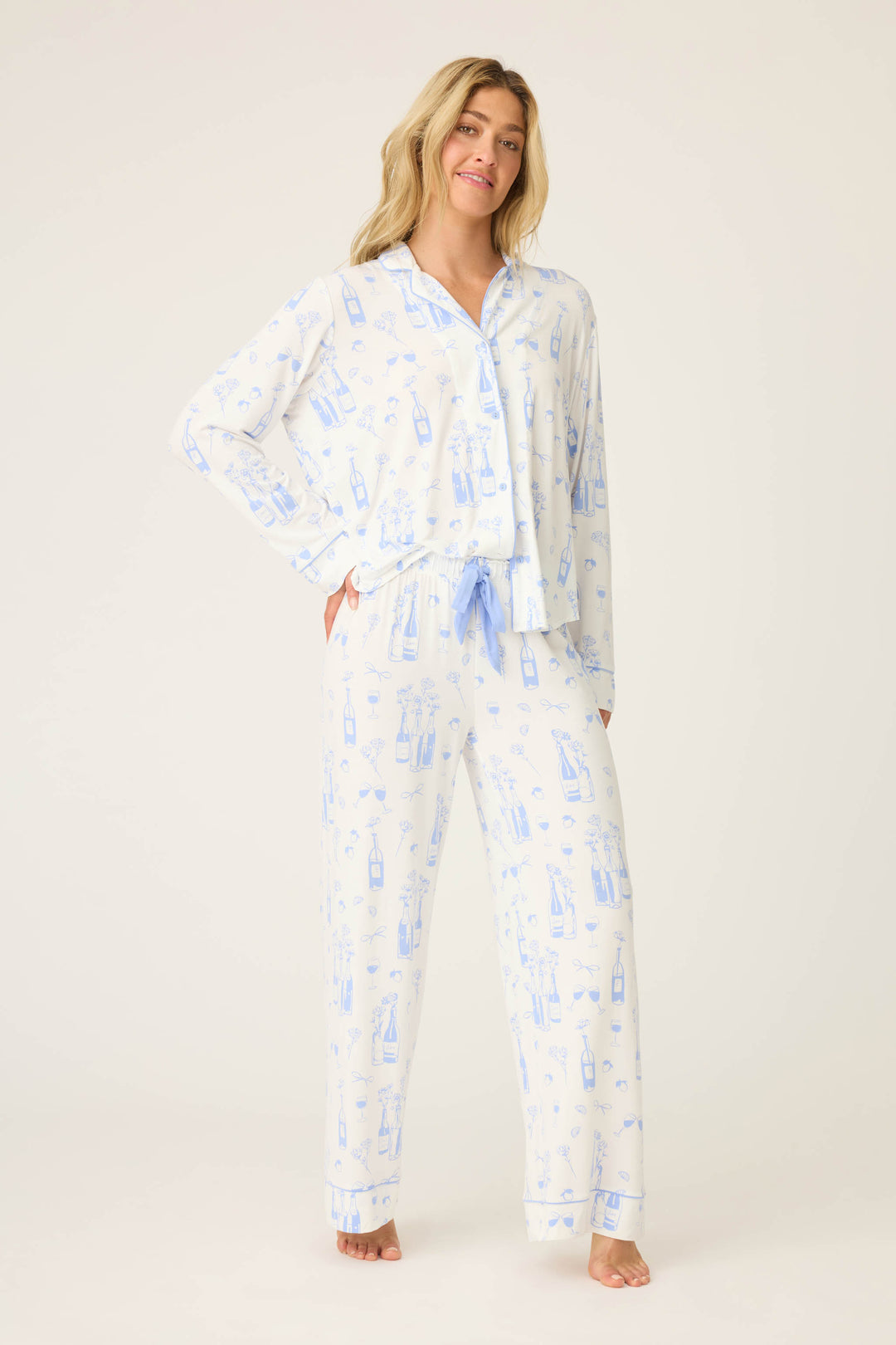 A woman stands barefoot against a plain background, wearing the P.J. Salvage Parisian Dreams Whimsical Print Open Leg Pant—a light blue and white pajama set featuring a wine bottle and glass print, with a button-up long sleeve top and drawstring pants.