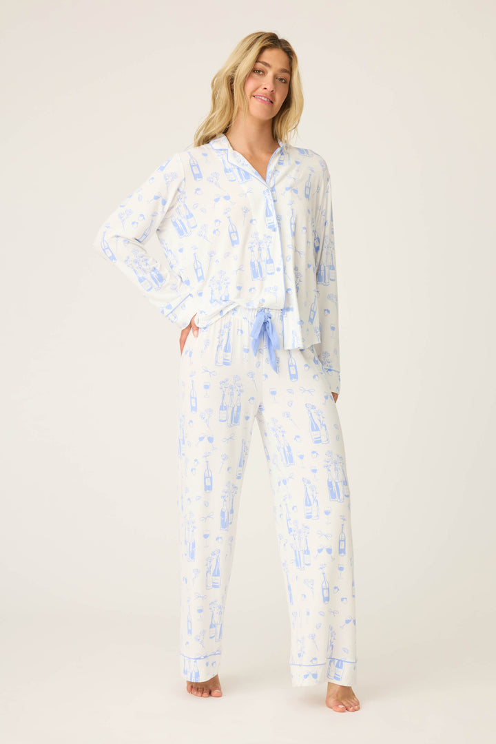 A woman stands barefoot against a plain background, wearing the P.J. Salvage Parisian Dreams Whimsical Print Open Leg Pant—a light blue and white pajama set featuring a wine bottle and glass print, with a button-up long sleeve top and drawstring pants.