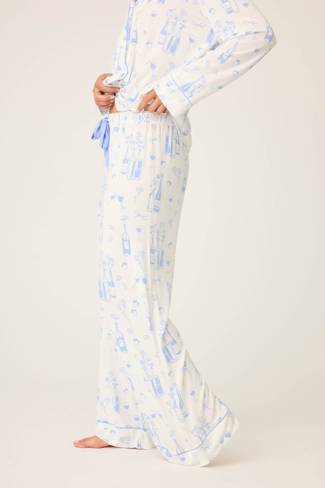 A person stands barefoot in light-colored, long-sleeved pajamas featuring P.J. Salvages Parisian Dreams Whimsical Print Open Leg Pant with a blue bottle and glass motif; their head is not visible in the image.