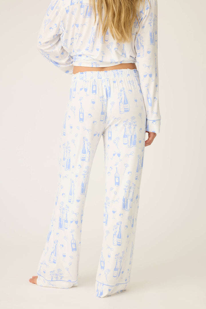A person stands barefoot facing away, wearing P.J. Salvage’s Parisian Dreams Whimsical Print Open Leg Pant with a blue bottle and glass pattern; only their lower back and legs are visible.