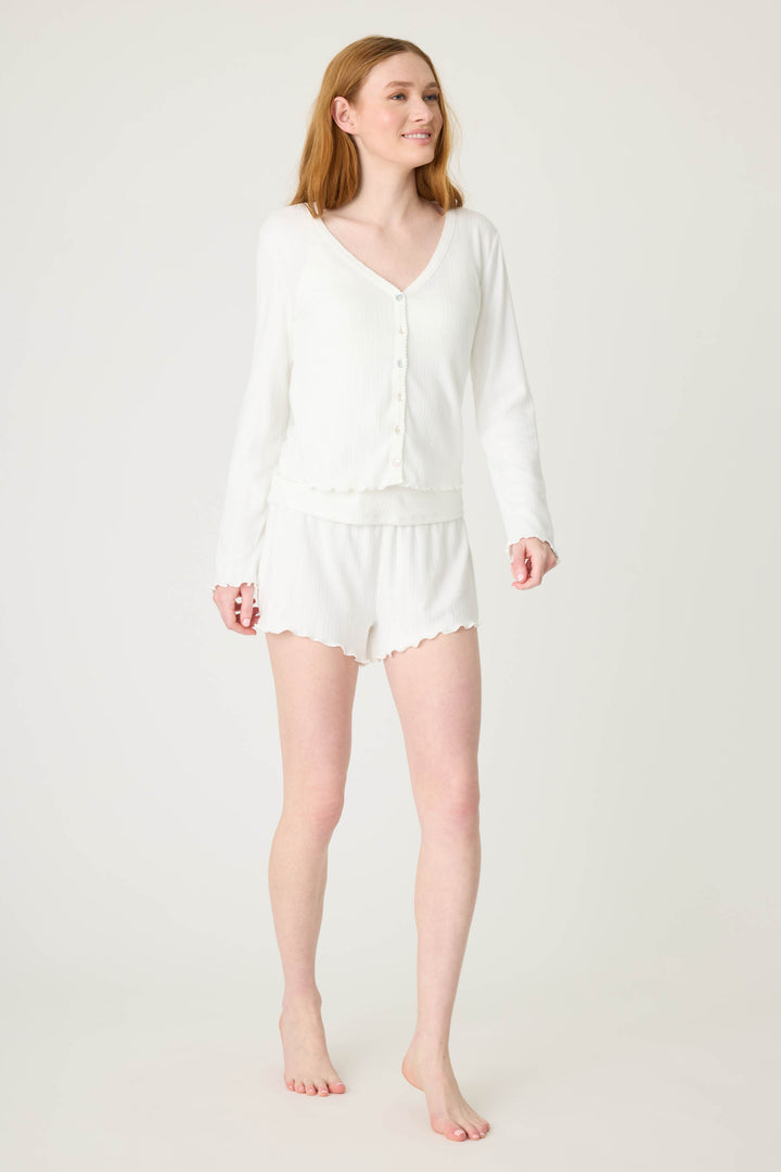 A woman with long red hair stands barefoot against a plain background, smiling slightly left while wearing the P.J. Salvage Pointelle Essentials Solid Ivory Cardigan with matching white shorts.