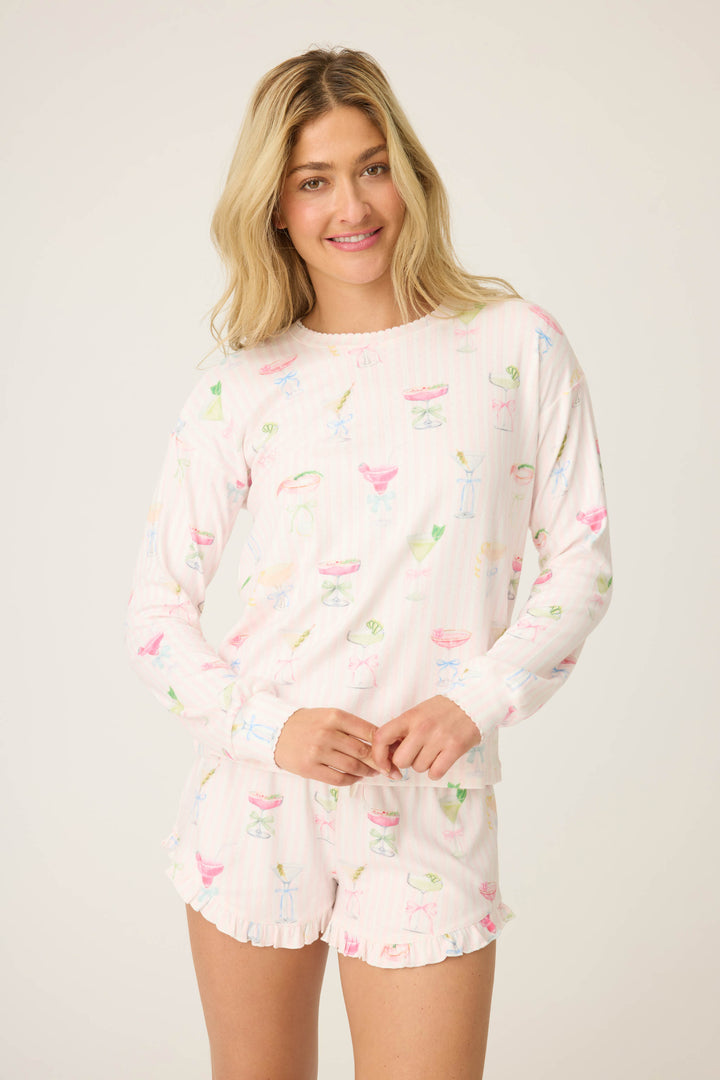 A woman with long blonde hair wears the Pretty Punch Cocktail Print Long Sleeve Top by P.J. Salvage—white with colorful cocktails—paired with ruffled shorts, smiling while standing against a plain background.