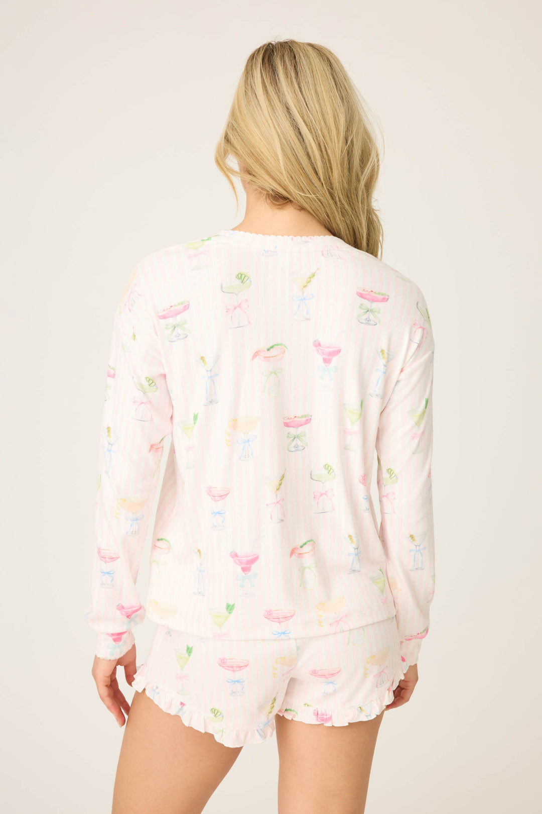 A woman with long blonde hair faces away from the camera, wearing the P.J. Salvage Pretty Punch Cocktail Print Long Sleeve Top and matching ruffled shorts. The pajama set features colorful patterns against a light background.