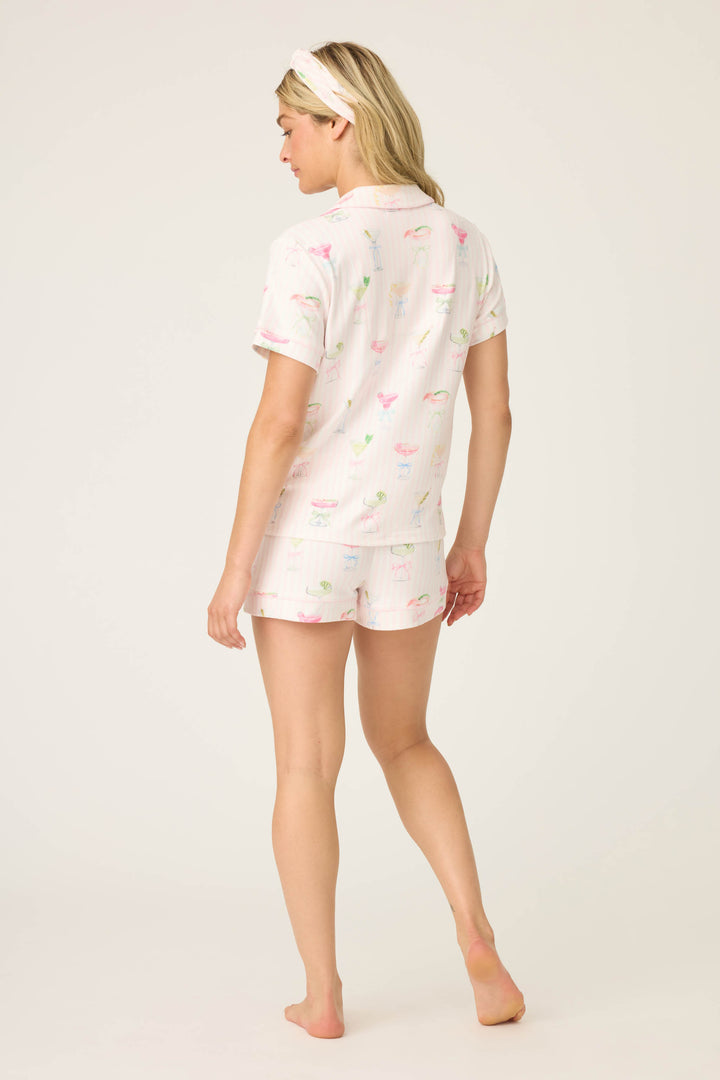 A woman stands barefoot facing away, wearing the P.J. Salvage Pretty Punch Cocktail Print 3-Piece Short Set with a pastel pattern and a light headband, against a plain background.