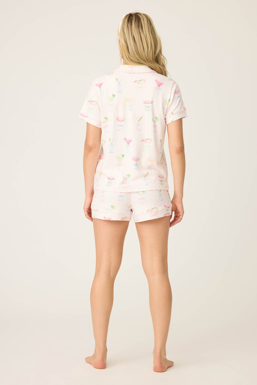 A woman with long blonde hair stands barefoot, facing away, wearing the P.J. Salvage Pretty Punch Cocktail Print 3-Piece Short Set, featuring a light color and colorful cocktail glass patterns, against a plain light background.