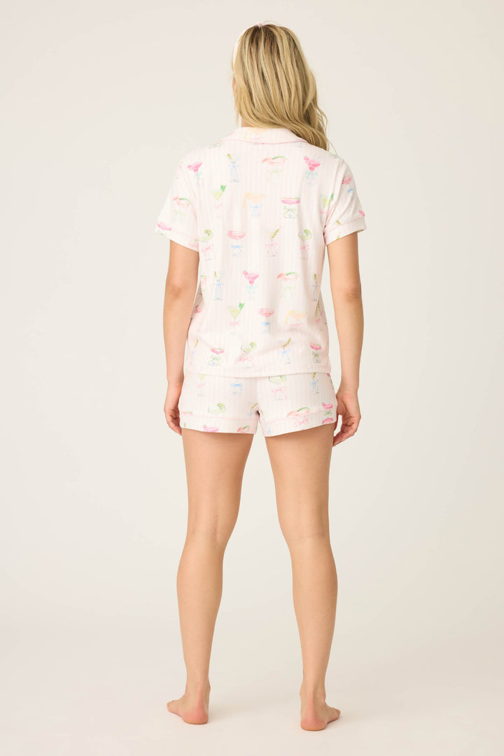 A woman with long blonde hair stands barefoot, facing away, wearing the P.J. Salvage Pretty Punch Cocktail Print 3-Piece Short Set, featuring a light color and colorful cocktail glass patterns, against a plain light background.