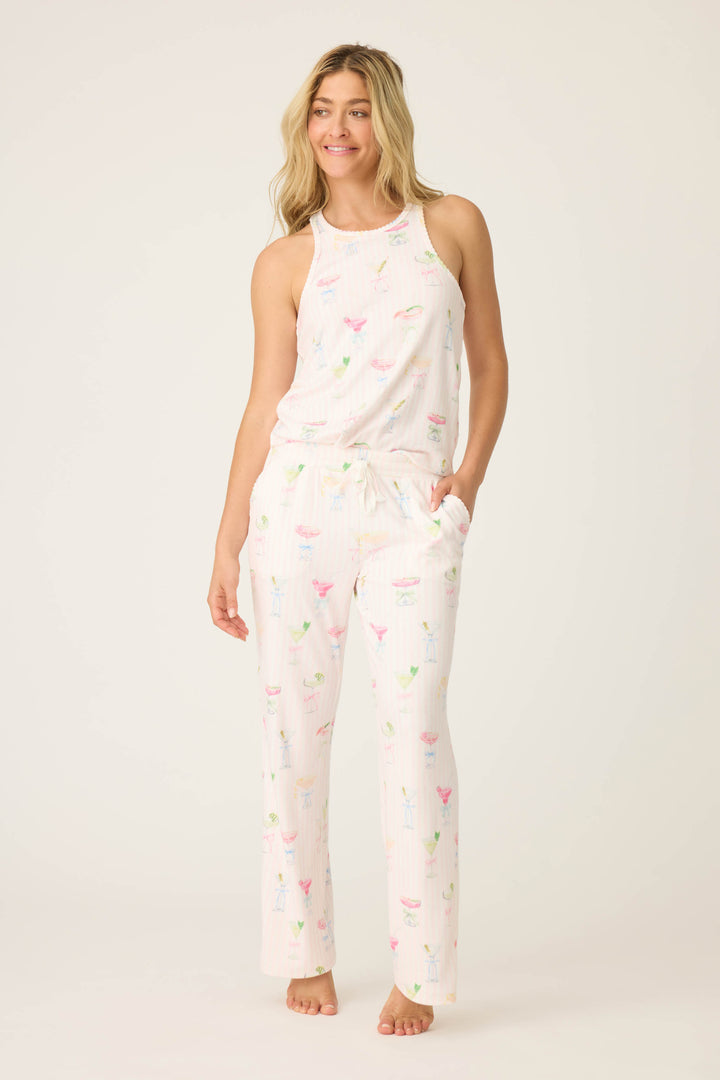 A woman with long blonde hair stands barefoot against a plain background, smiling slightly and wearing P.J. Salvages Pretty Punch Cocktail Print Open Leg Pants, with a white base and colorful cocktail glass pattern. One hand is in her pocket.