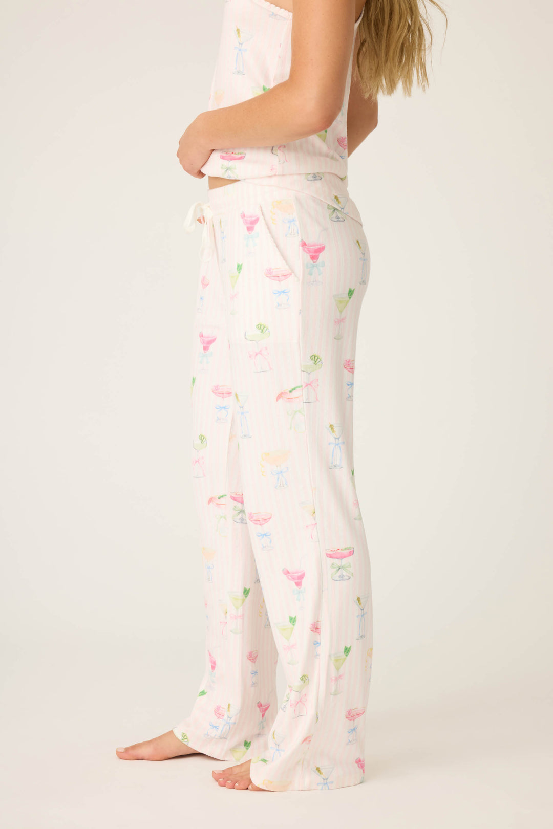 A person stands barefoot, shown from the upper chest down, wearing P.J. Salvage Pretty Punch Cocktail Print Open Leg Pants—light-colored pajama pants with a drawstring waist and a pastel cocktail glass pattern.