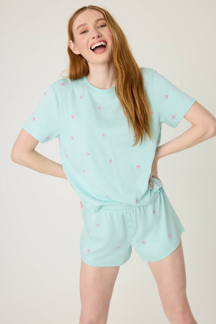 A person with long red hair smiles with hands on hips, wearing the P.J. Salvage Pretty Punch Bow Print Loungewear Short Set—light blue short sleeves and matching shorts with small pink designs—against a plain light background.