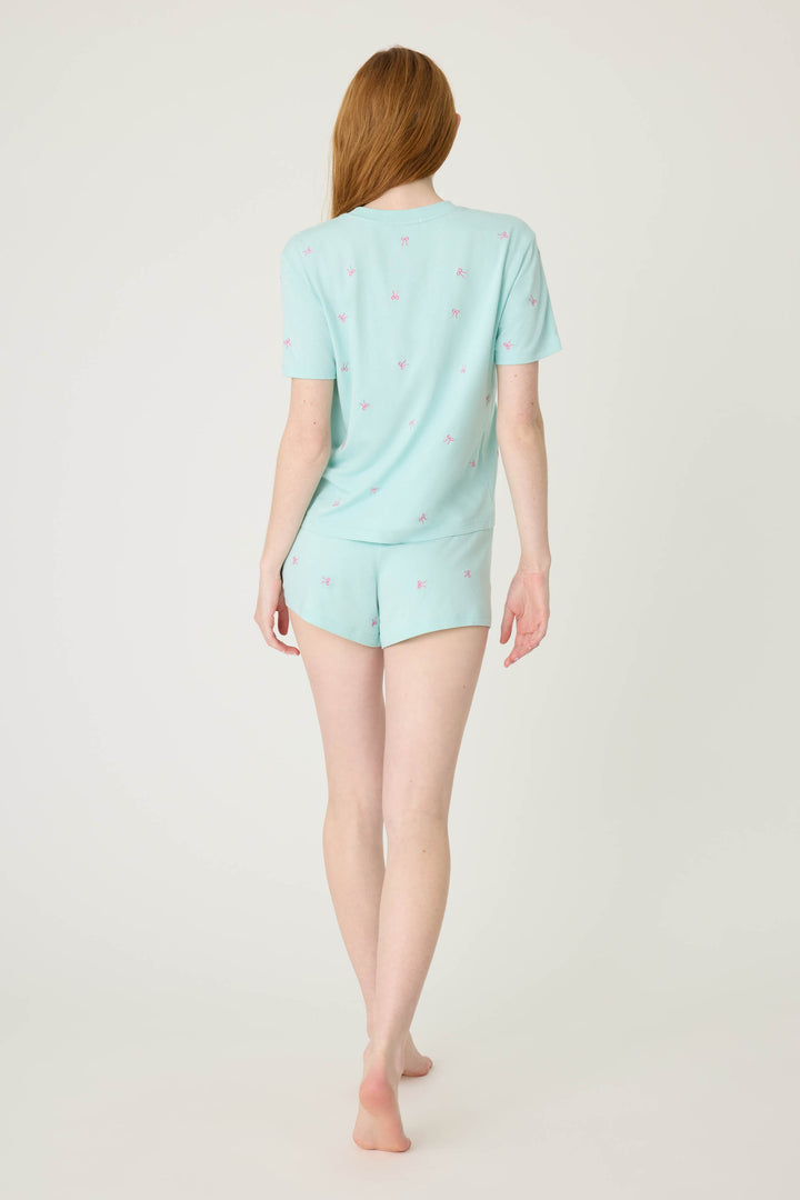 A woman with long red hair stands barefoot facing away, wearing the P.J. Salvage Pretty Punch Bow Print Loungewear Short Set—a matching light blue short-sleeve pajama set with a subtle bow pattern—against a plain light background.