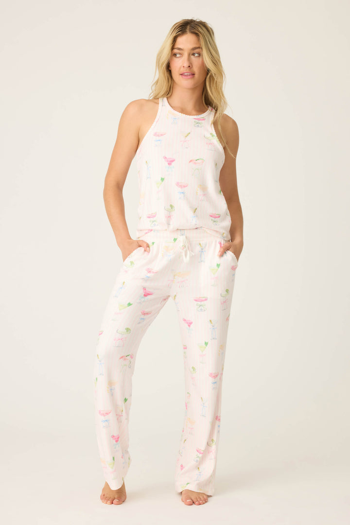 A person stands barefoot against a plain background wearing the Pretty Punch Cocktail Print PJ Set by P.J. Salvage, featuring a white sleeveless tank and matching drawstring pajama pants with colorful cocktail glass prints. Hands are in the pockets; hair is loose.