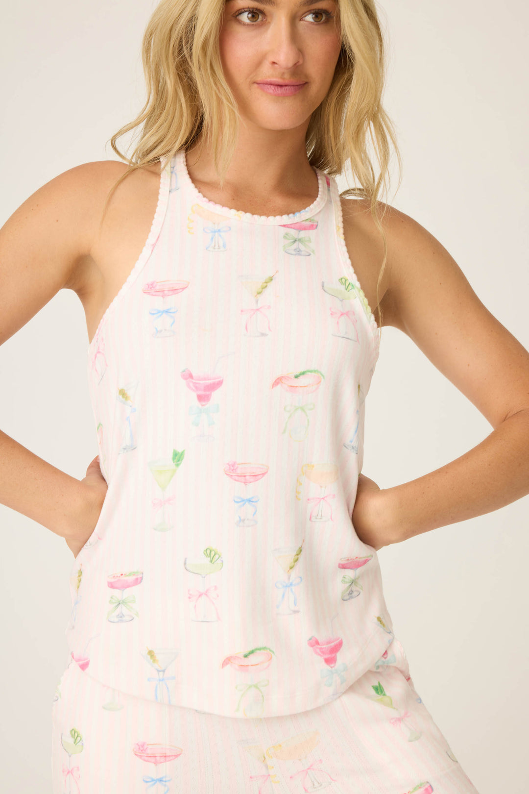 A woman wears the P.J. Salvage Pretty Punch Cocktail Print PJ Set—a white sleeveless top with light pink stripes and colorful cocktail glass designs—standing with her hands on her hips, looking slightly off camera.