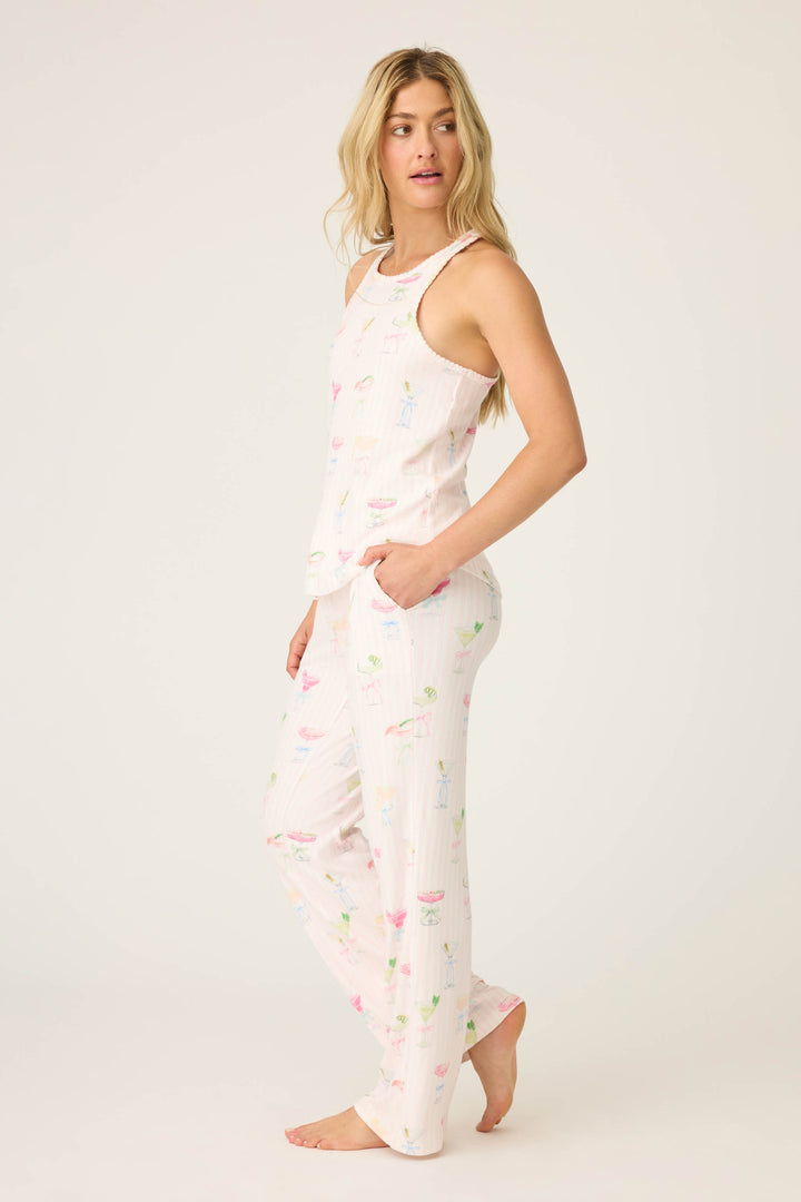 A woman with long, loose hair stands barefoot against a plain background in the P.J. Salvage Pretty Punch Cocktail Print PJ Set—a matching white tank top and loose pants decorated with colorful drink and umbrella patterns, one hand tucked in her pocket.
