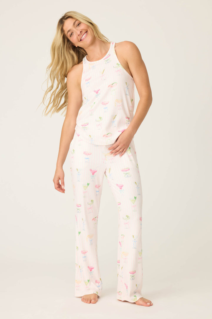 A woman with long blonde hair smiles barefoot in the P.J. Salvage Pretty Punch Cocktail Print PJ Set—a matching light pink, sleeveless top and wide-leg pants with a colorful drink pattern—against a plain light background.