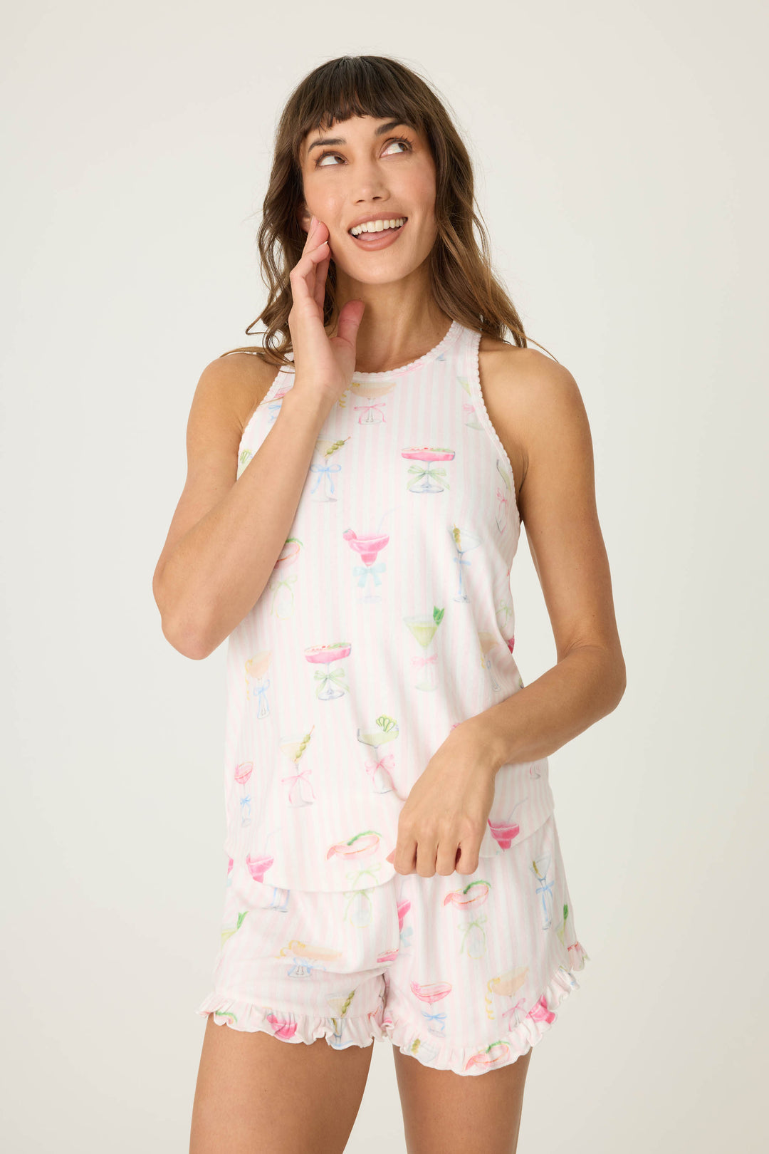 A woman stands against a plain background, smiling and touching her face, wearing the P.J. Salvage Pretty Punch Cocktail Print Racerback Tank Top with matching shorts.