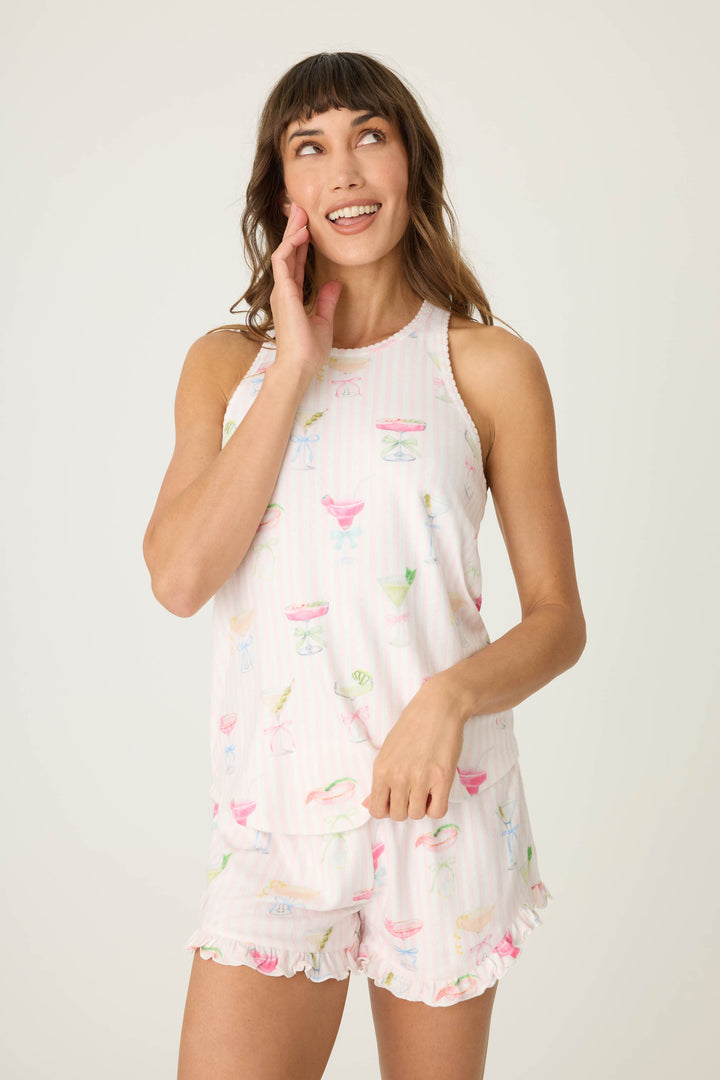 A woman stands against a plain background, smiling and touching her face, wearing the P.J. Salvage Pretty Punch Cocktail Print Racerback Tank Top with matching shorts.