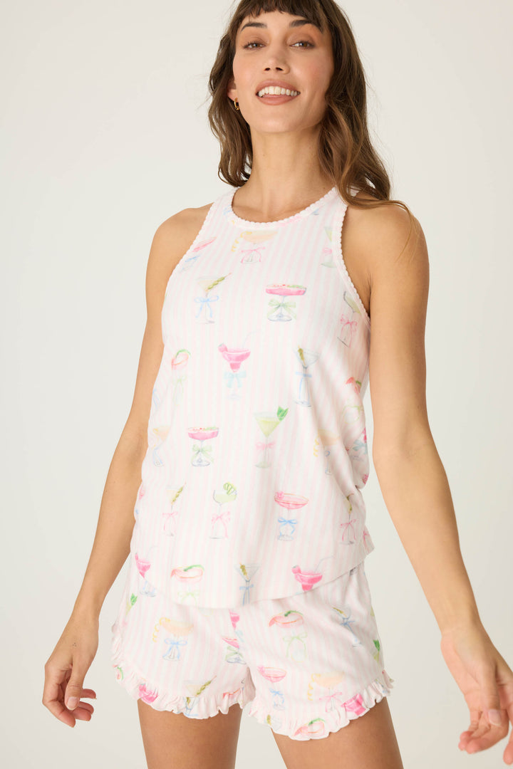 A woman smiles in a P.J. Salvage Pretty Punch Cocktail Print Racerback Tank Top with ruffled shorts, featuring a fun light pink cocktail glass pattern against a plain, light background.