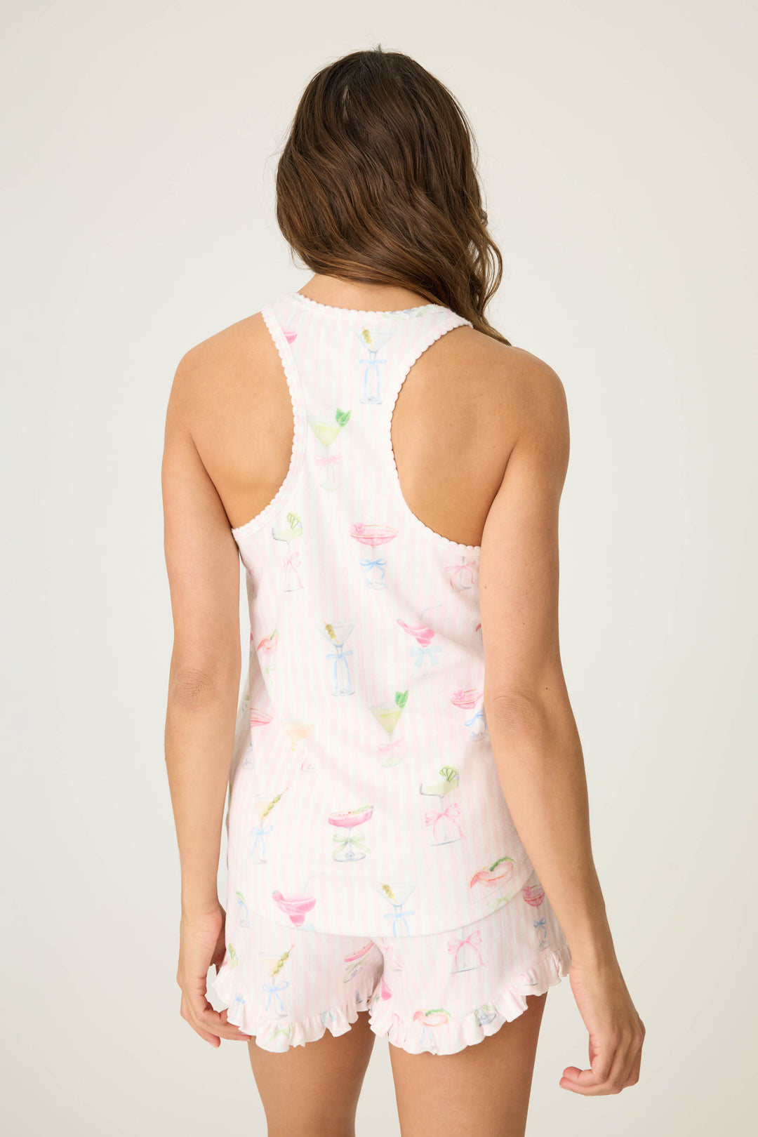 A woman with long brown hair faces away, modeling the P.J. Salvage Pretty Punch Cocktail Print Racerback Tank Top in light pink with pastel drink prints and a ruffled hem. The background is plain light beige.