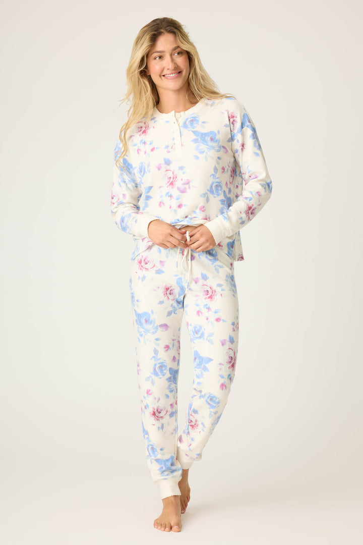 A person models the P.J. Salvage Pant Set FLORAL PJ SET RODEO ROSE, featuring matching long sleeves with blue and pink floral patterns on a white background, standing barefoot against a plain light backdrop and smiling to the side.