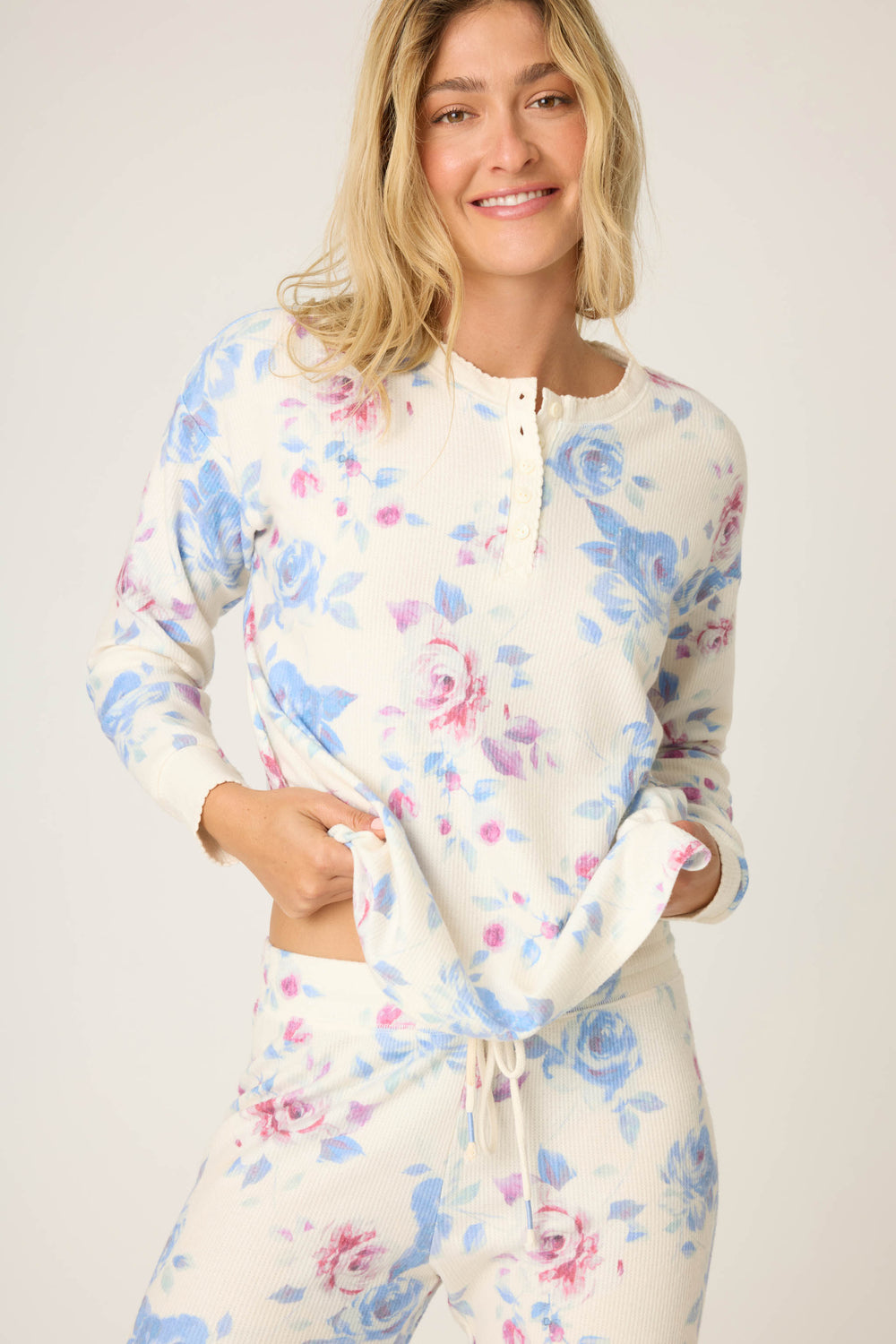 A person wearing the P.J. Salvage Pant Set FLORAL PJ SET RODEO ROSE, featuring a blue and pink floral print top and pants, stands against a light background, smiling and holding the hem of the shirt.