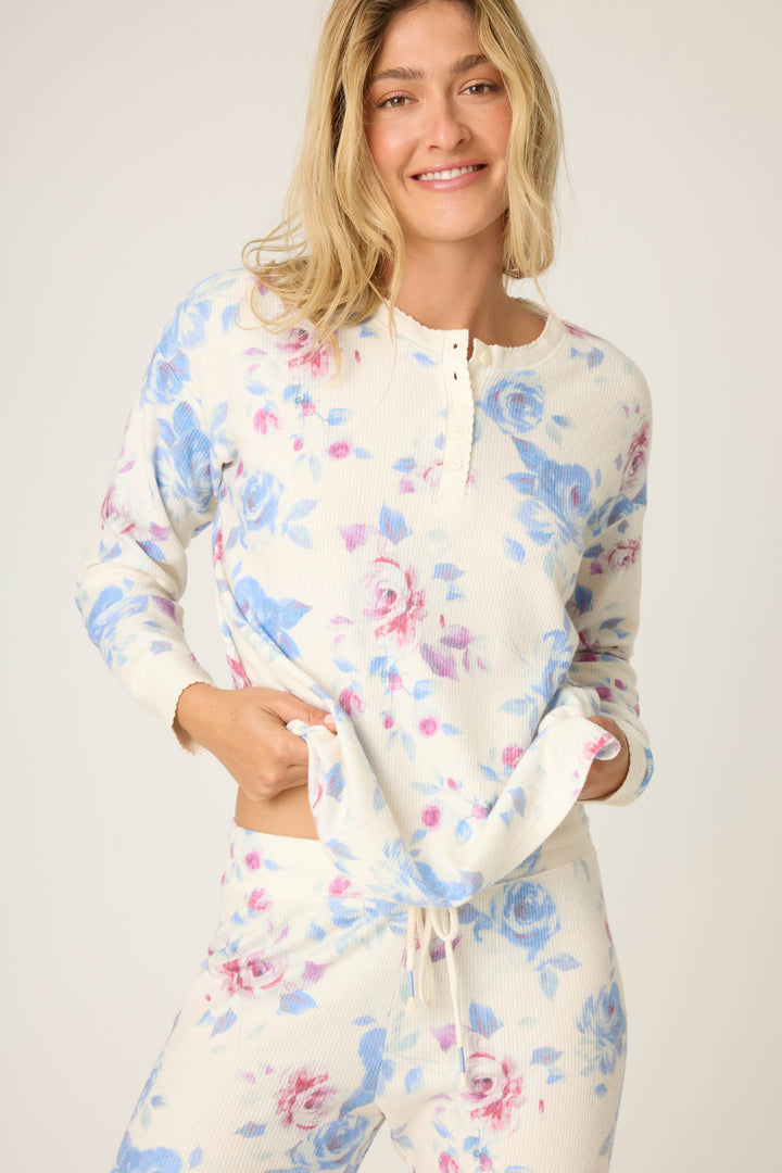 A person wearing the P.J. Salvage Pant Set FLORAL PJ SET RODEO ROSE, featuring a blue and pink floral print top and pants, stands against a light background, smiling and holding the hem of the shirt.