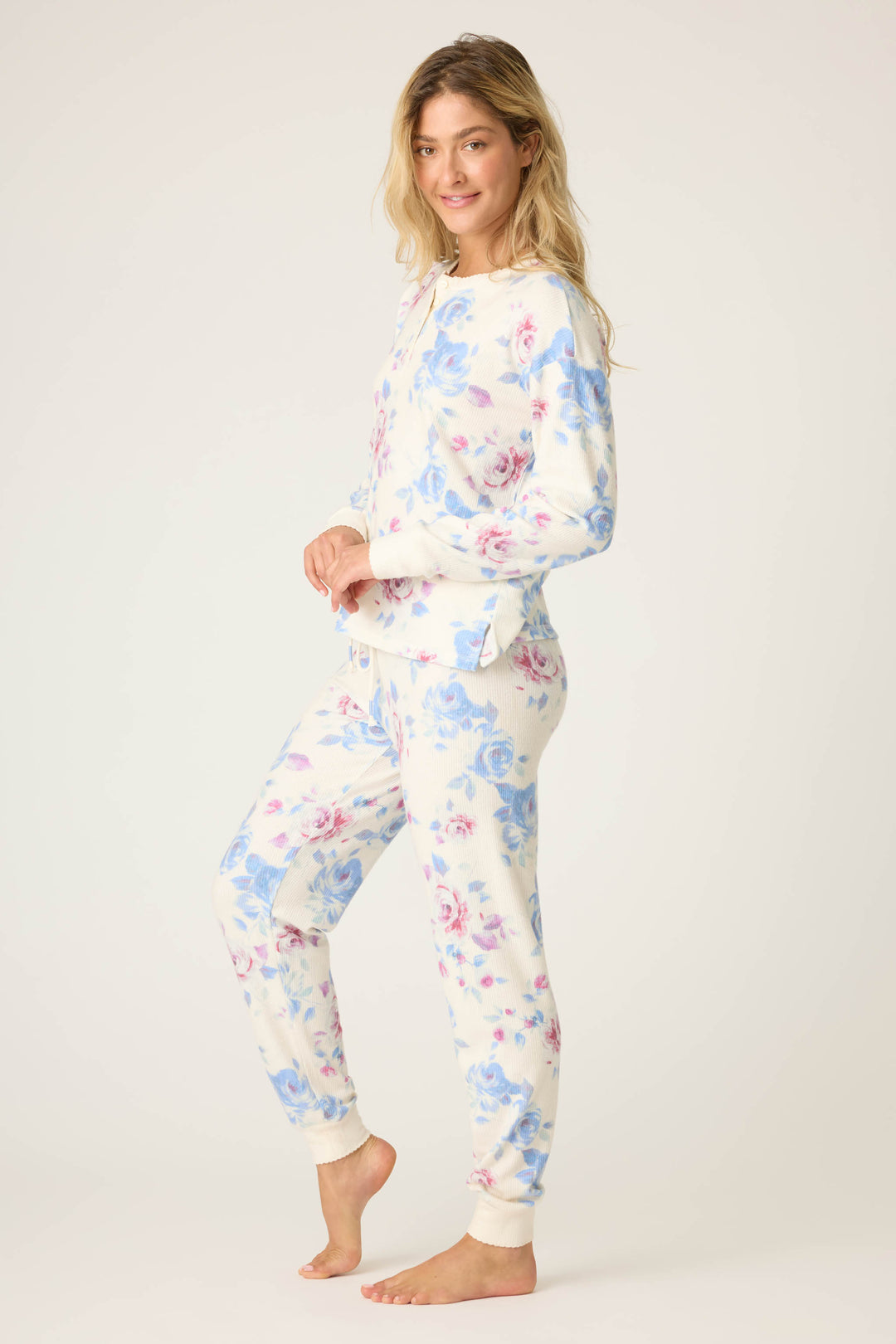 A person stands barefoot against a plain background, wearing the P.J. Salvage Pant Set FLORAL PJ SET RODEO ROSE—matching long-sleeve pajamas with a white base and blue and pink floral print—looking at the camera with a slight smile.