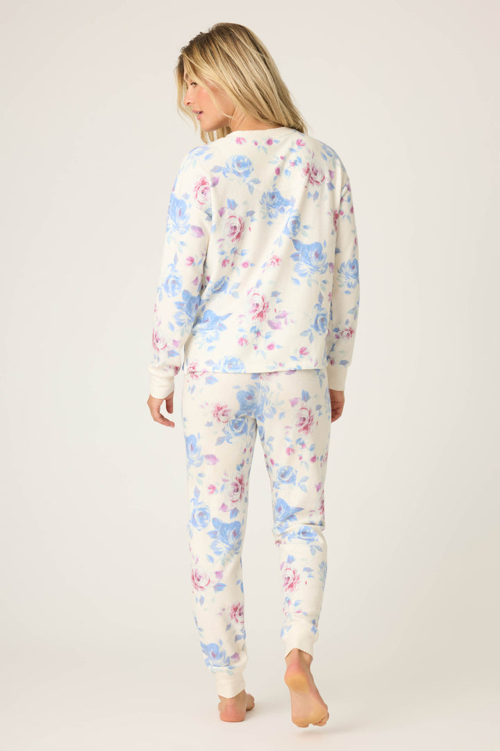 A person with long blonde hair stands barefoot, facing away, wearing the P.J. Salvage Pant Set FLORAL PJ SET RODEO ROSE—a matching long-sleeve top and pants with a white background and blue and pink floral pattern. Background is plain and light-colored.