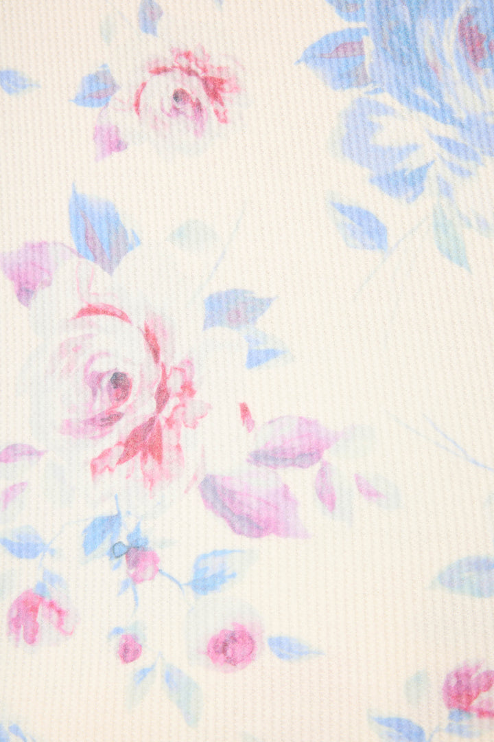 A close-up of the P.J. Salvage Pant Set FLORAL PJ SET RODEO ROSE shows light pink and blue roses with leaves on a cream, slightly ribbed fabric.