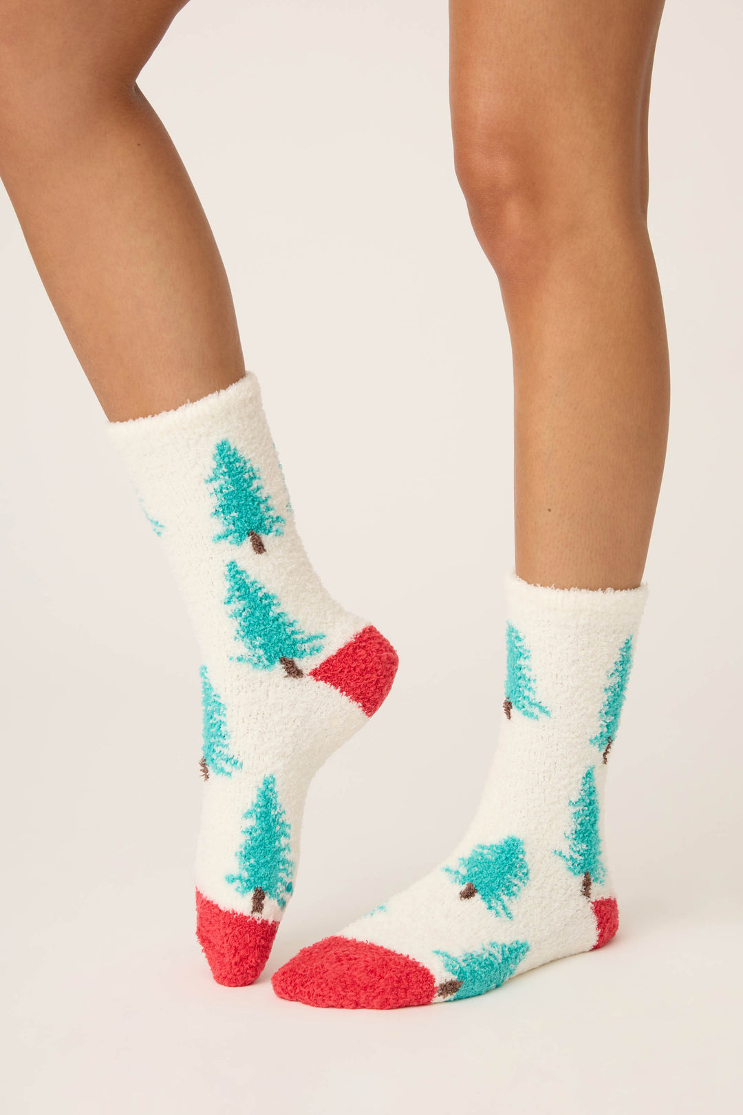 A person wears P.J. Salvage Winter Tree Fuzzy Crew Socks with green Christmas tree patterns and red heels and toes. Only their lower legs and feet are visible against a plain, light-colored background.