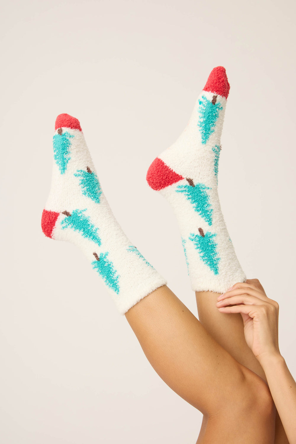 Two legs in the air wear P.J. Salvage Winter Tree Fuzzy Crew Socks—white with red toes and heels, decorated with teal Christmas trees. One hand gently holds an ankle against a plain, light background.