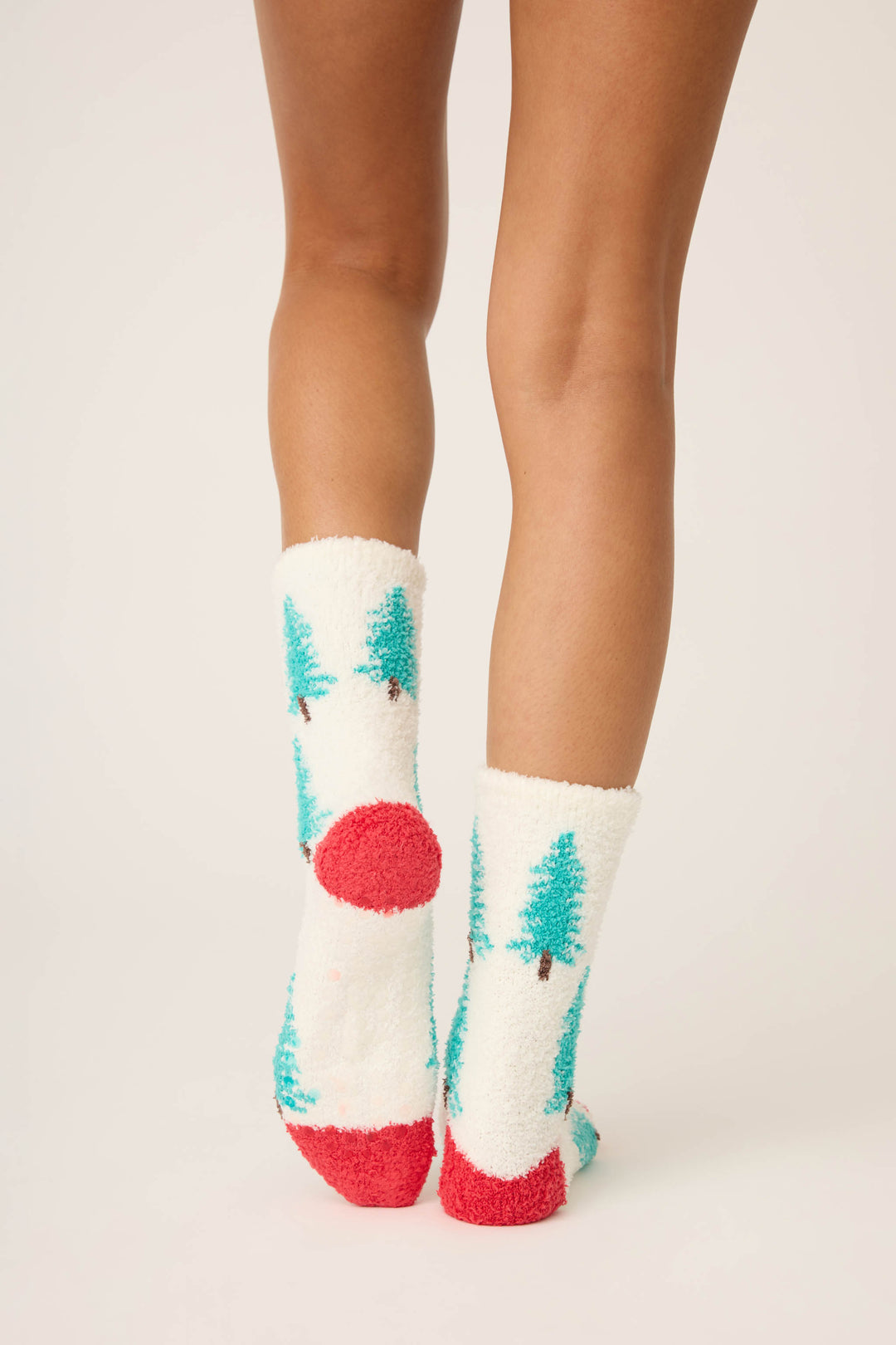 A person stands with ankles crossed, wearing P.J. Salvage Winter Tree Fuzzy Crew Socks—white socks with green trees and red accents on the toes, heels, and soles. The background is plain and light-colored.