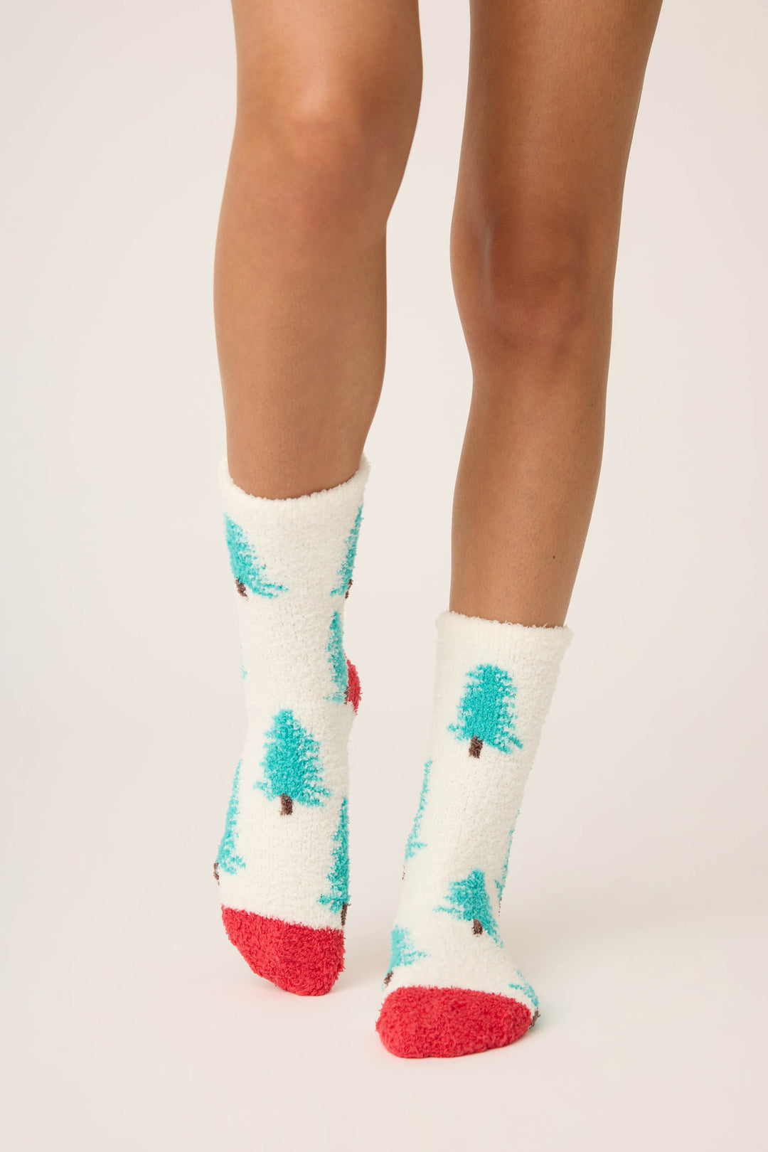 A person models P.J. Salvage Winter Tree Fuzzy Crew Socks: cozy white socks with blue-green tree patterns, red toes and heels. Only their legs and feet are shown against a plain, light background.