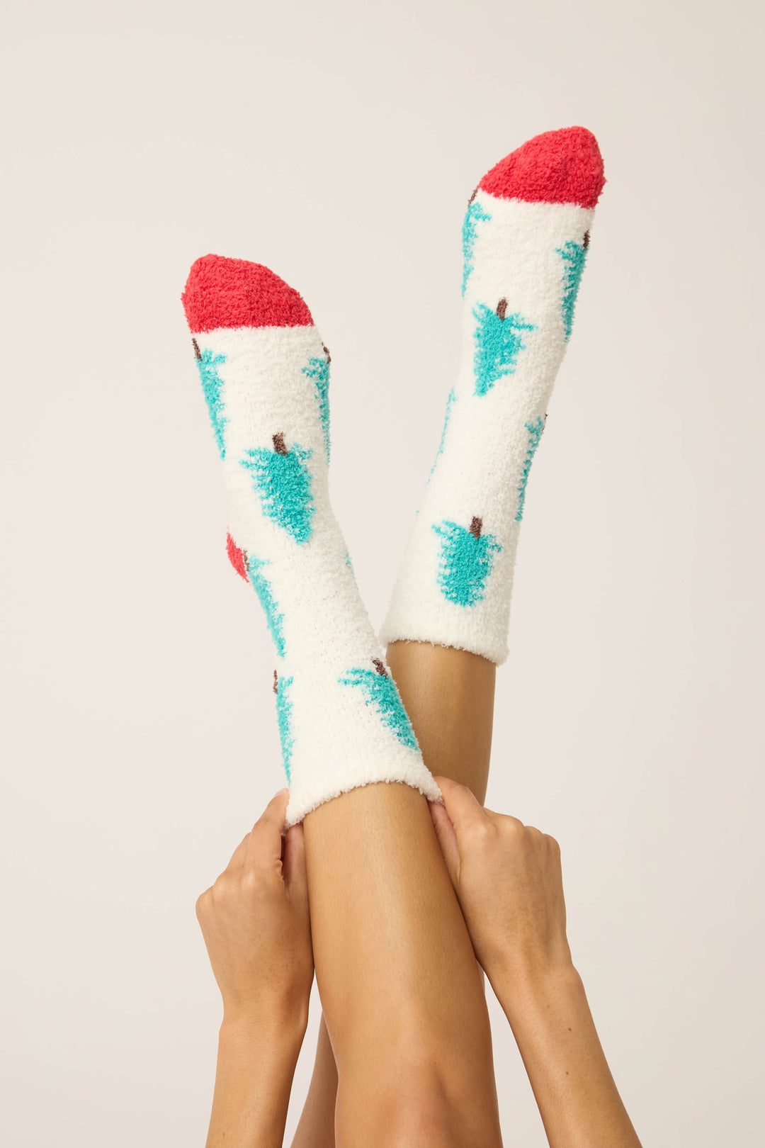 A pair of crossed legs showcase P.J. Salvage’s Winter Tree Fuzzy Crew Socks—white with teal Christmas tree patterns and red toe caps—against a simple, light background.