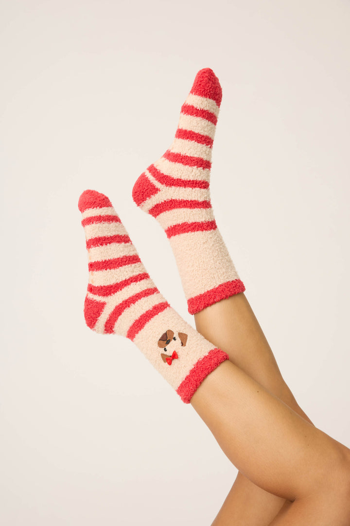 A person lies on a plain white background with legs raised, wearing P.J. Salvage Striped Dog Fuzzy Crew Socks—red and white stripes, plus a small dog face with a red bow tie near the ankle.