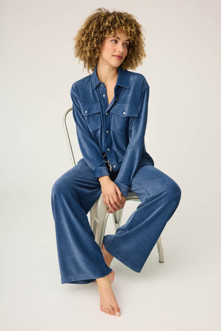 A person with curly hair sits barefoot on a metal chair against a plain background, wearing the Craving Corduroy Velour Lounge Set by P.J. Salvage—a blue retro-inspired long-sleeve shirt and matching wide-leg pants.