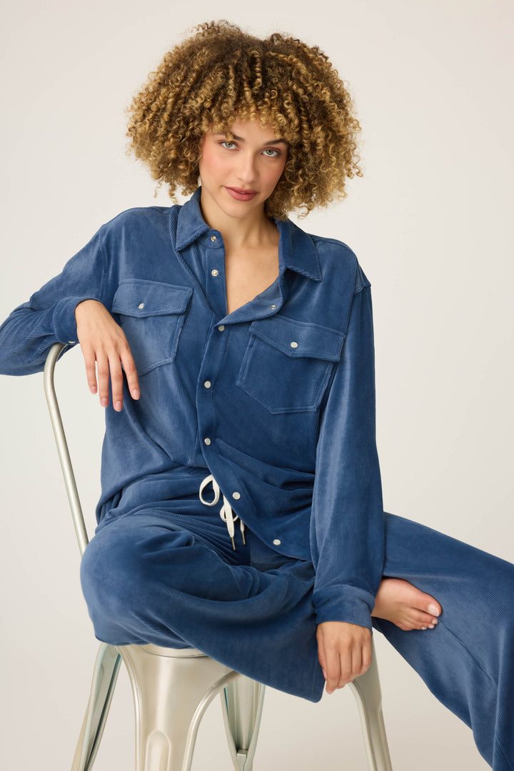 A person with curly hair sits sideways on a metal chair wearing P.J. Salvage’s Craving Corduroy Velour Lounge Set—an oversized blue retro button-up top with large pockets and matching wide-leg pants—against a plain background.