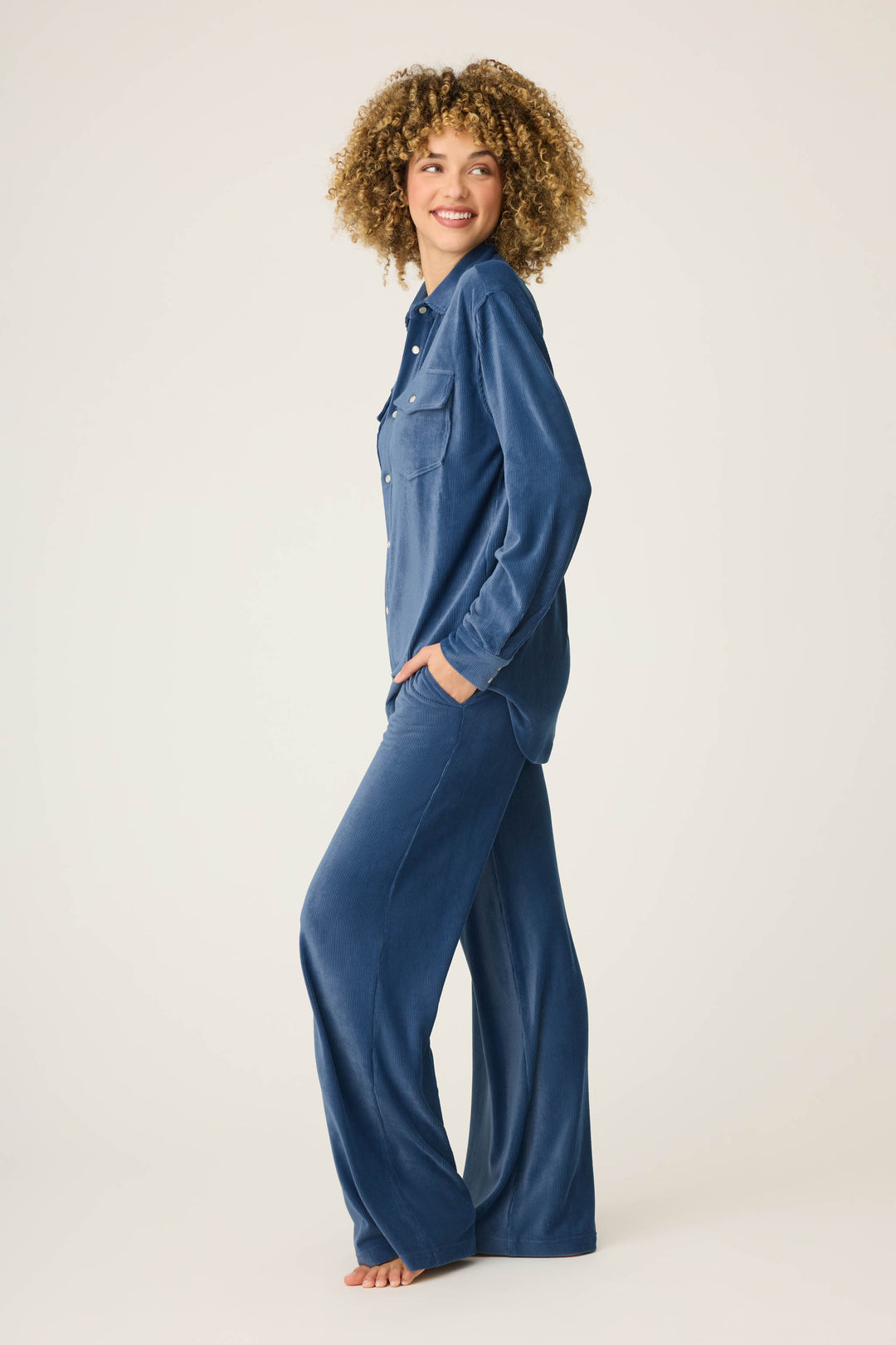A person with curly hair models the P.J. Salvage Craving Corduroy Velour Lounge Set, featuring retro button-up top and wide-leg pants, standing barefoot on a plain background with hands in pockets, smiling and looking to the side.
