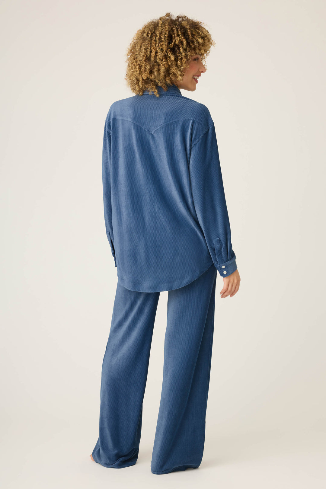 Model with curly hair stands barefoot and faces away, wearing the P.J. Salvage Craving Corduroy Velour Lounge Set—featuring a loose retro button-up top and matching wide-leg pants—against a plain light background.