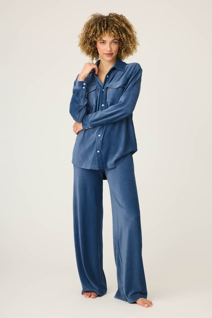 A person with curly hair models the P.J. Salvage Craving Corduroy Velour Lounge Set—a blue retro button-up shirt with chest pockets and matching wide-leg pants—standing barefoot against a plain light background.