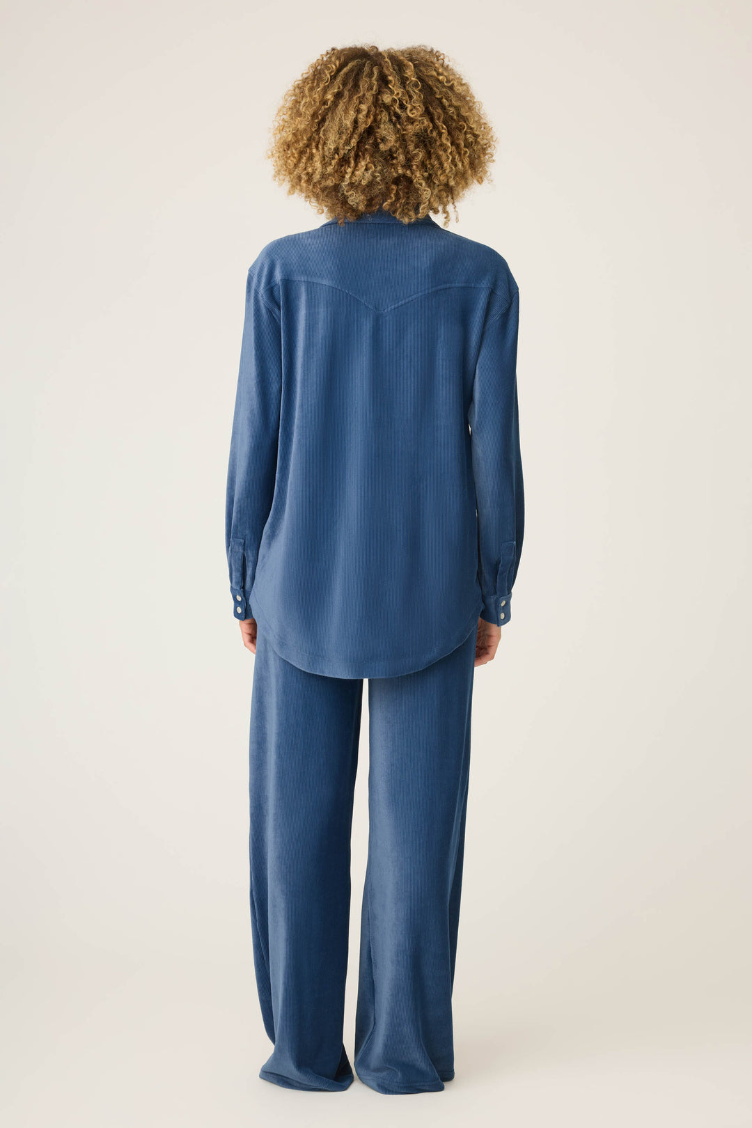 A person with curly hair stands facing away, wearing the P.J. Salvage Craving Corduroy Velour Lounge Set—a loose-fitting, long-sleeve button-up shirt and matching wide-leg blue pants—against a plain light background.