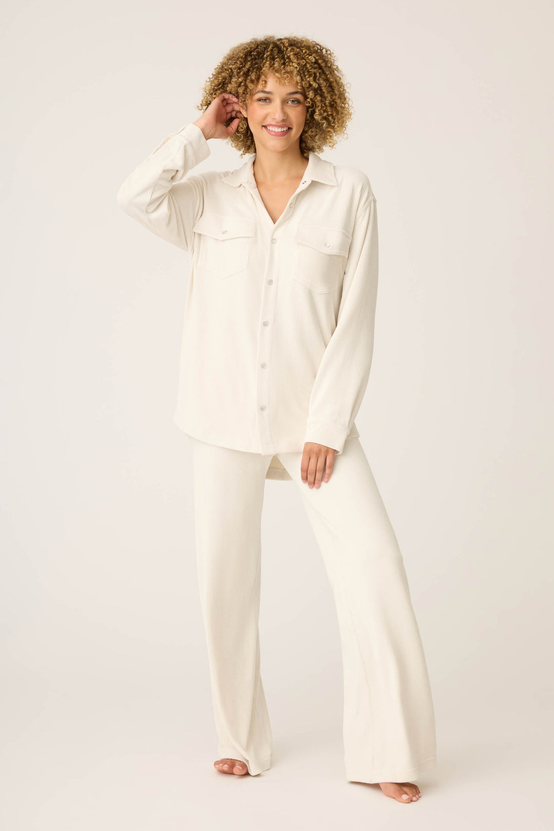 A person stands barefoot on a plain background, smiling and touching their head, wearing the SOLID PJ SET CRAVING CORDUROY by P.J. Salvage—an off-white button-up set with front pockets and matching wide-leg pants.