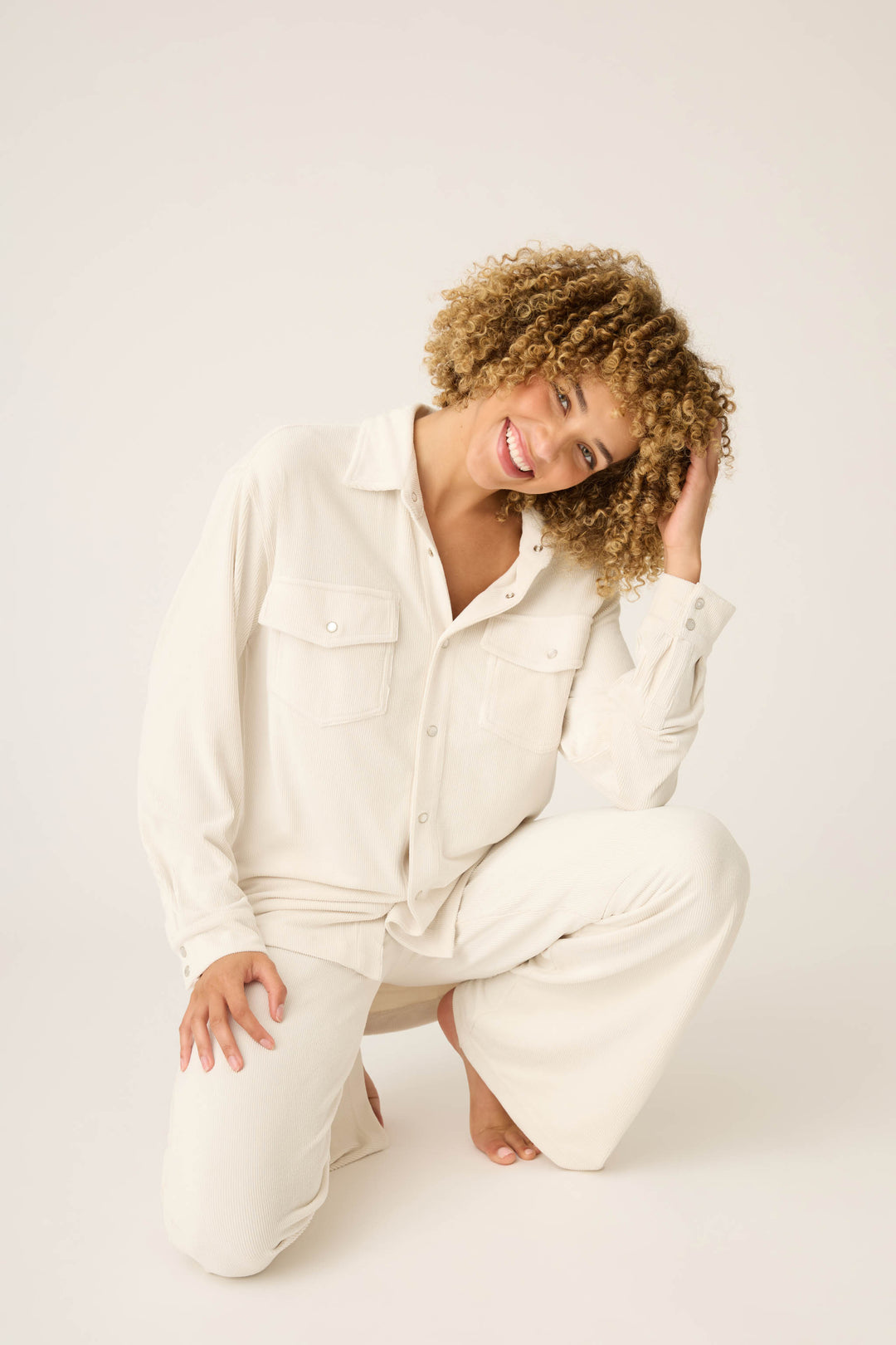 A person with curly hair is smiling and kneeling on one knee, touching their hair, while wearing the SOLID PJ SET CRAVING CORDUROY by P.J. Salvage against a plain background.