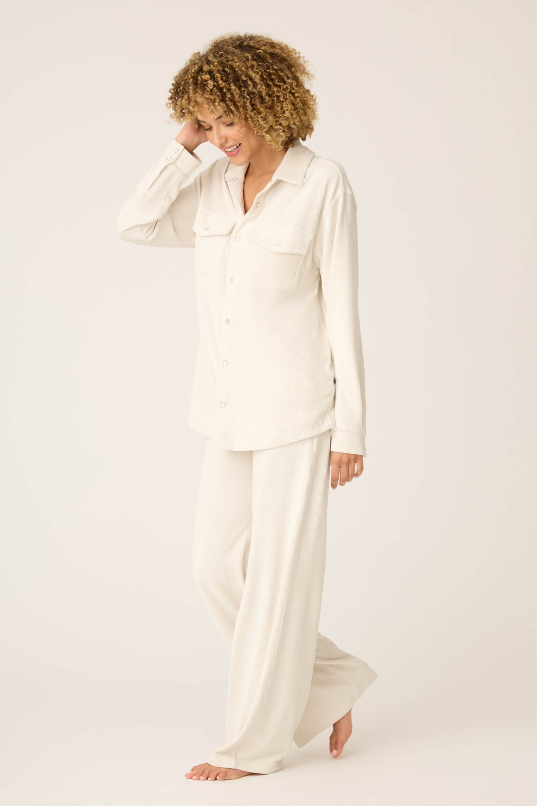 A person with curly hair stands barefoot against a plain background, wearing the SOLID PJ SET CRAVING CORDUROY by P.J. Salvage—featuring a loose, long-sleeved cream button-up shirt and matching wide-leg pants.