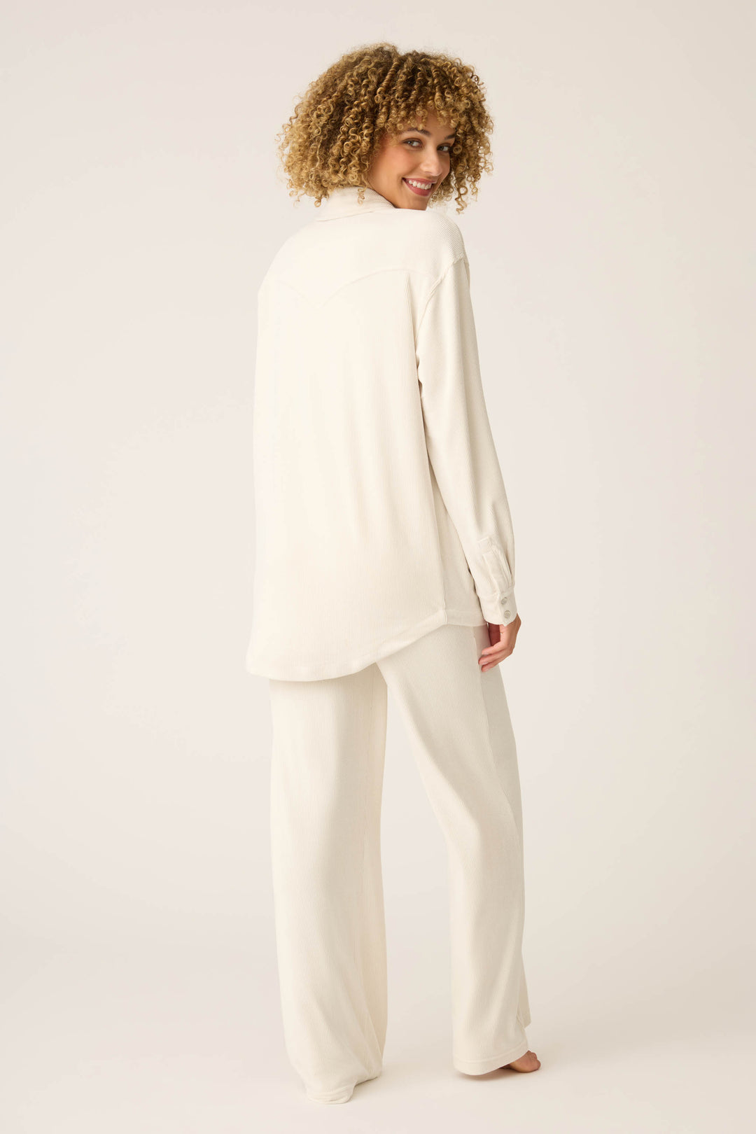 A person with curly hair stands barefoot against a plain background, wearing the SOLID PJ SET CRAVING CORDUROY by P.J. Salvage—a loose, long-sleeved cream top and matching wide-leg pants—while looking over their shoulder.