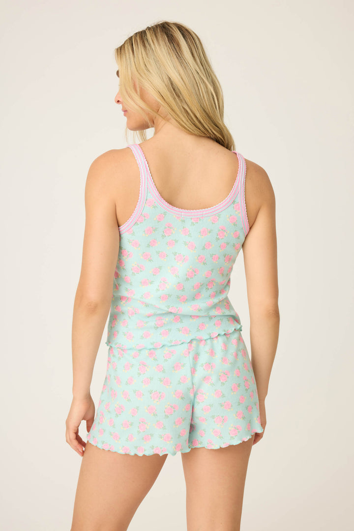 A person with long blonde hair is seen from behind wearing the P.J. Salvage Secret Garden Floral Camisole set—mint green with a pink floral pattern and scalloped edges. The tank top and shorts are shown against a plain, light-colored background.